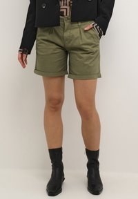 Shorts en coton vert olive avec un ourlet roulé, comportant des plis et une fermeture à bouton à l'avant. Assortis à une veste noire et des bottines en cuir.