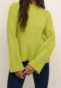 Pull vert vif à côtes avec de larges manches évasées et un col montant. Le tissu semble doux avec une texture tricotée épaisse ; l'ourlet est légèrement raccourci.