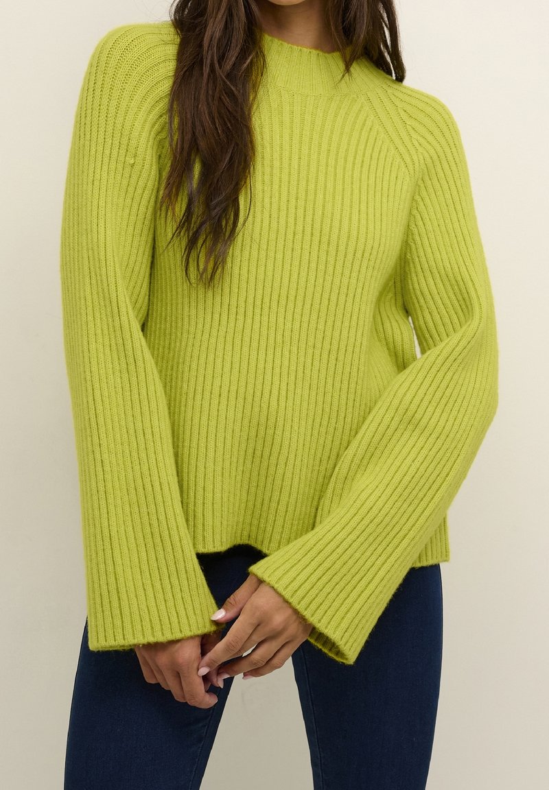 Pull vert vif à côtes avec de larges manches évasées et un col montant. Le tissu semble doux avec une texture tricotée épaisse ; l'ourlet est légèrement raccourci.