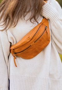 Osoba u kremastom pletenom puloveru nosi svijetlosmeđu kožnu torbicu preko tijela s više zatvorenih odjeljaka.