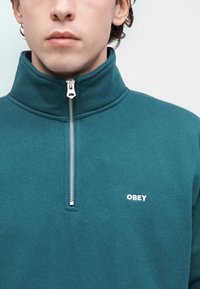 Φούτερ σε τυρκουάζ με φερμουάρ, κατασκευασμένο από μαλακό ύφασμα. Διαθέτει ψηλό γιακά και ασημένιο φερμουάρ, με μικρό λευκό λογότυπο "OBEY" στο στήθος.