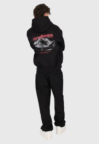 Person in einem schwarzen Hoodie mit einem Bergmotiv und dem Text "endless", schwarzen Hosen und weißen Turnschuhen, steht mit dem Rücken zur Kamera.