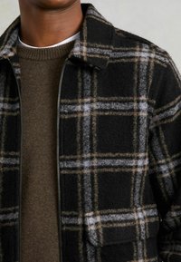 Sort og ternet jakke med en uldne tekstur, der har grå, hvid og tan striber samt en frontlynlås. Brun sweater under.
