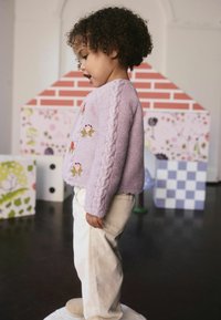 Peuter in een lavendel trui met bloemgeborduurde details en lichte broek staat op een witte ondergrond in een kamer met kleurrijke blokken.