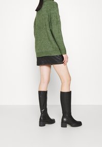 Pull en maille vert avec un motif texturé, associé à une mini jupe noire matelassée et des bottes en cuir noires à talons carrés.