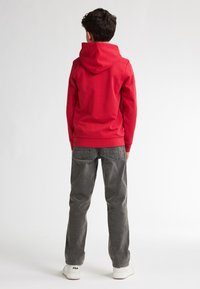 Felpa rossa con cappuccio e tasca a kangaroo, abbinata a jeans in denim grigi. Modello che indossa sneaker bianche con il marchio visibile.