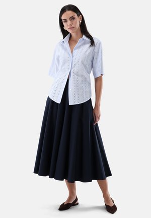 Woman wearing light blue eyelet short-sleeve shirt paired with a high-waisted, full, dark navy midi skirt and brown pointed flats.