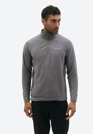 Mees, kes kannab helehallit kvartetizuuga fliisipulloverit ja musti pükse, vaatab küljele, seistes ühetaolise heleda tausta ees.