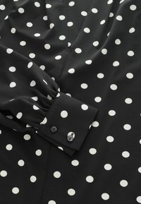 Fekete anyag fehér pöttyökkel, mély kivágással és ráncolt mandzsettákkal, az ujján két hozzáillő gombbal díszítve.