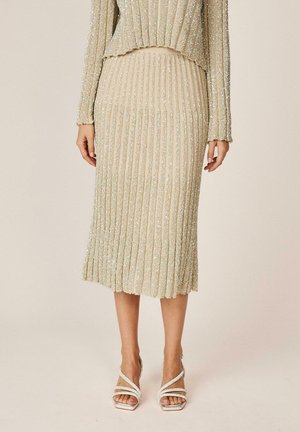 Falda midi plisada con brillo dorado combinada con top de manga larga a juego y sandalias de tacón alto con tiras sobre fondo neutro.