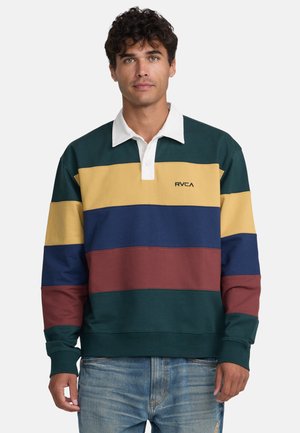 Jonge man draagt een meerkleurig gestreept rugbyshirt met groene, gele, blauwe en bordeauxrode banen en lichtblauwe spijkerbroek, staand tegen een effen achtergrond.
