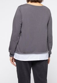 Sudadera gris con mangas largas y escote redondo, superpuesta sobre una camisa blanca. Presenta un ajuste relajado y puños acanalados.