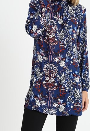 Blauwe tuniek met lange mouwen en ingewikkelde bloemen- en wijnrankpatronen in wit, rood en roze, gedragen over een zwarte broek.