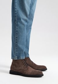 Brązowe zamszowe botki za kostkę z czarnymi sznurówkami, okrągłym czubkiem i czarną gumową podeszwą, zestawione z jasnoniebieskimi dżinsami.