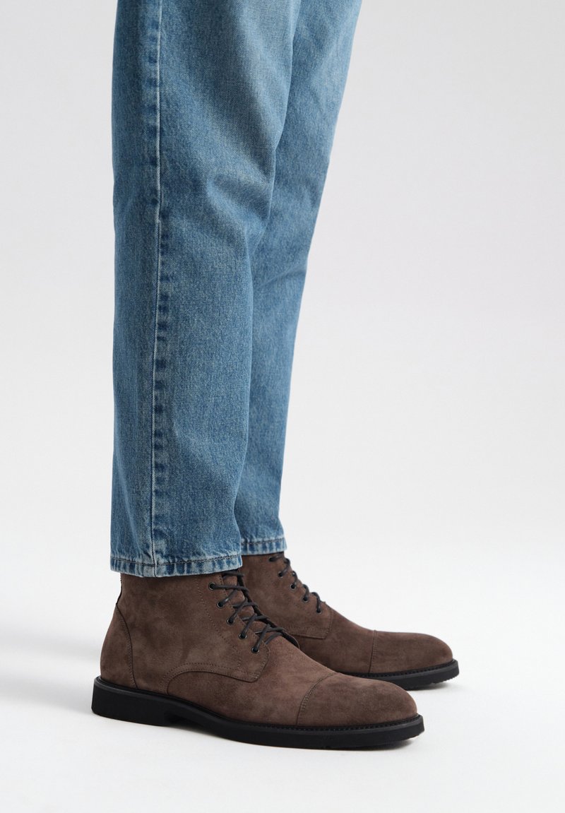 Brązowe zamszowe botki za kostkę z czarnymi sznurówkami, okrągłym czubkiem i czarną gumową podeszwą, zestawione z jasnoniebieskimi dżinsami.