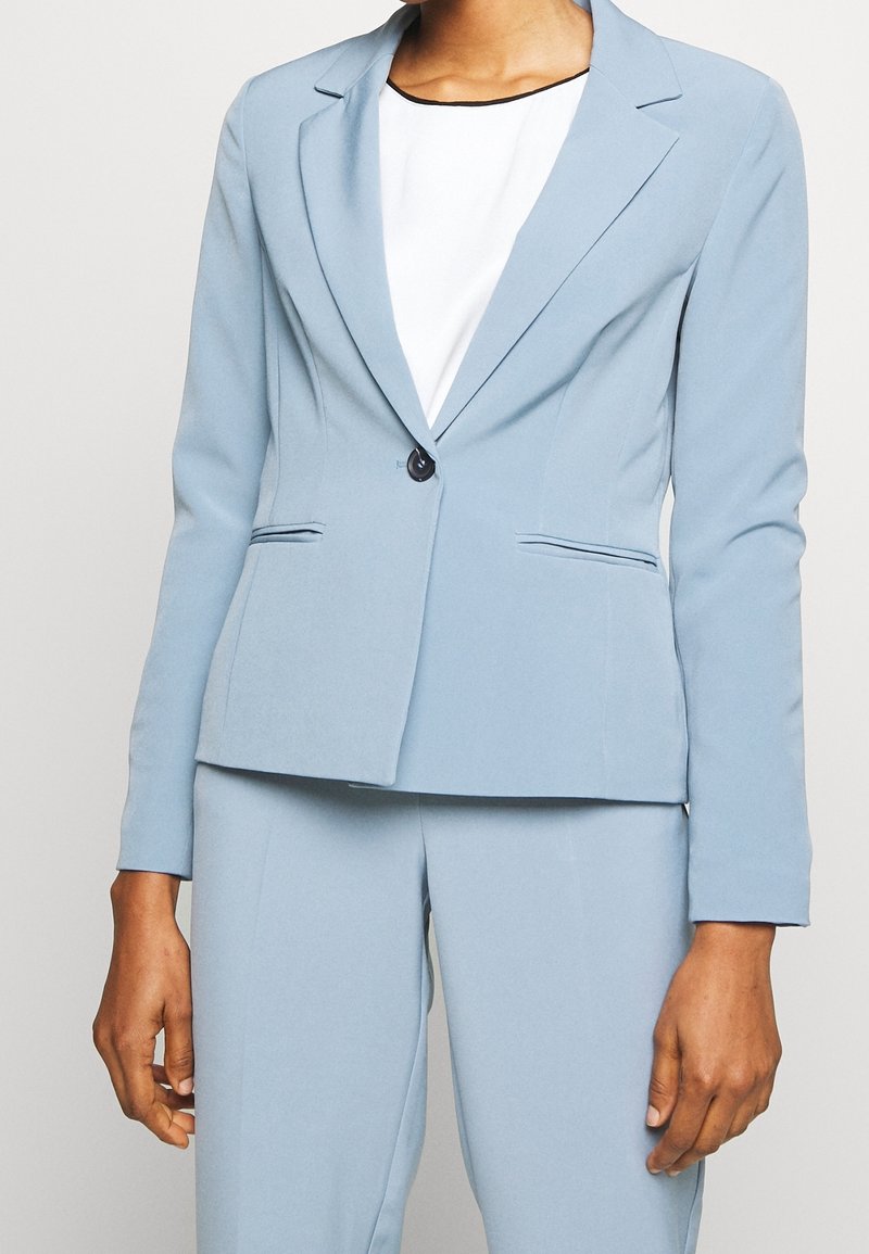 Blazer bleu clair slim fit avec un seul bouton noir sur un top blanc, assorti à un pantalon bleu clair.