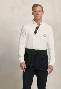 Cămașă polo albă cu mâneci lungi și buzunar, logo de tenis verde, asortată cu pantalonii bleumarin cu dungi fine și un pulover tricotat verde închis legat în talie.