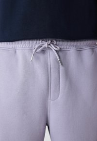 Pantalon de survêtement violet clair avec une taille élastique et un cordon de serrage noué, porté avec un haut sombre, montré de la taille à mi-cuisse.