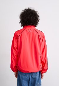 Red windbreaker jacket with black accents, high collar, and elastic cuffs. Features a logo on the upper back. Lightweight, smooth texture.