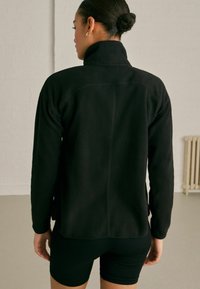 Giacca in pile nera con colletto alto, finitura testurizzata e design aderente. La vista posteriore mostra i dettagli della cucitura della spalla e le maniche lunghe.