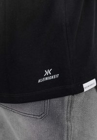 T-shirt noir avec logo "KLEINIGKEIT" près de l'ourlet, associé à un pantalon gris texturé montrant les coutures de la poche arrière.