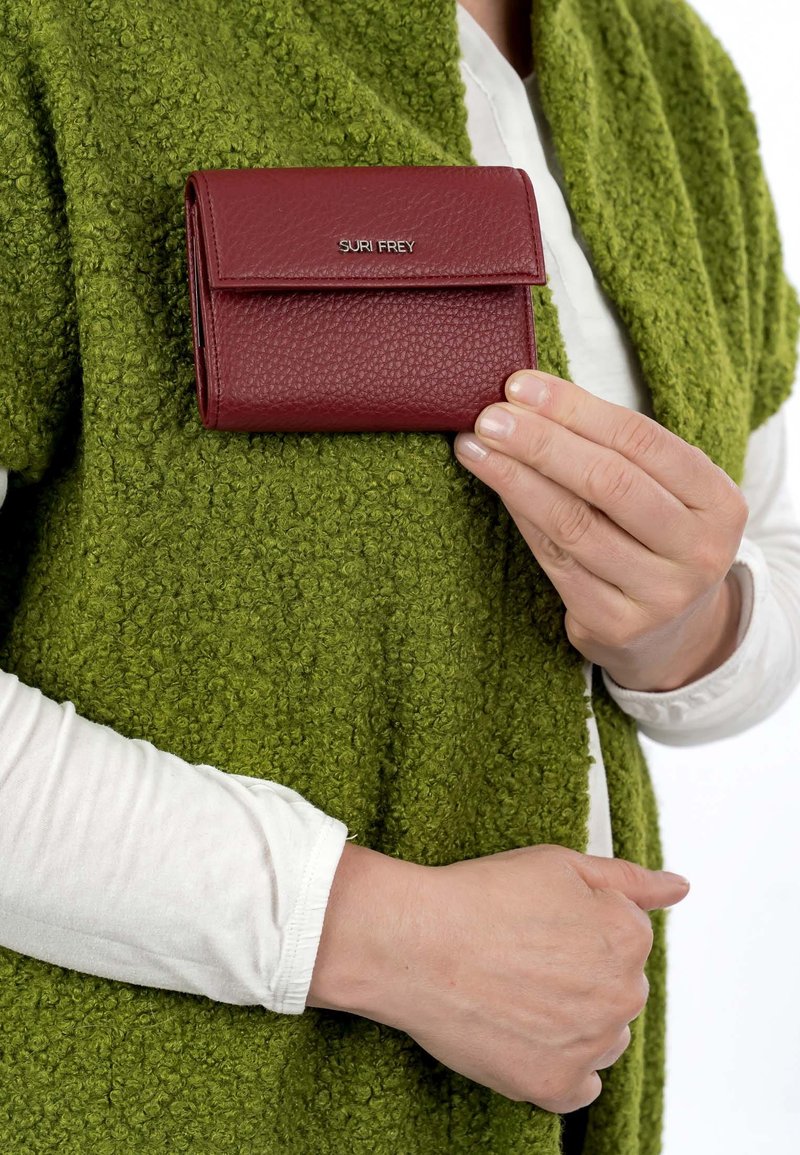 Personne tenant un portefeuille Suri Frey rouge texturé au-dessus d'un vêtement vert texturé, portant une chemise blanche à manches longues en dessous.