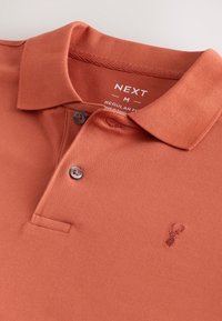 Polo in roestkleur met een gestructureerde stof, twee grijze knopen en een klein geborduurd logo op de borst. De halslijn heeft een klassieke kraag.