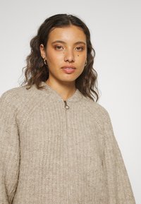 Beige gebreide jack met ritssluiting en ribbelstructuur, rond de hals. Voorzien van een zilveren ritssluiting en brede mouwen.