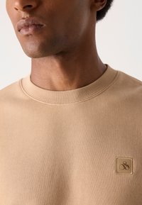 Sweatshirt en coton beige avec un col rond côtelé, featuring un patch de logo carré texturé sur la poitrine gauche. Texture de tissu lisse.