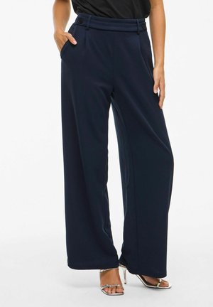 Vrouw in een losse, marineblauwe broek met zijzakken en zilveren hoge hakken, tegen een effen achtergrond.