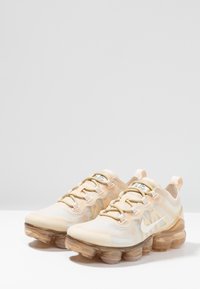 Baskets de sport beiges avec un dessus en mesh, un logo Nike discret et une semelle à air transparente et multicouche. Lacets plats et languettes pour un enfilage facile.