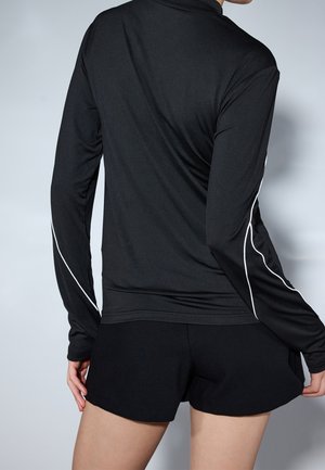 Kvinde iført sort atletisk langærmet top med hvid piping på ærmerne og sorte shorts, vist bagfra mod en ensfarvet baggrund.