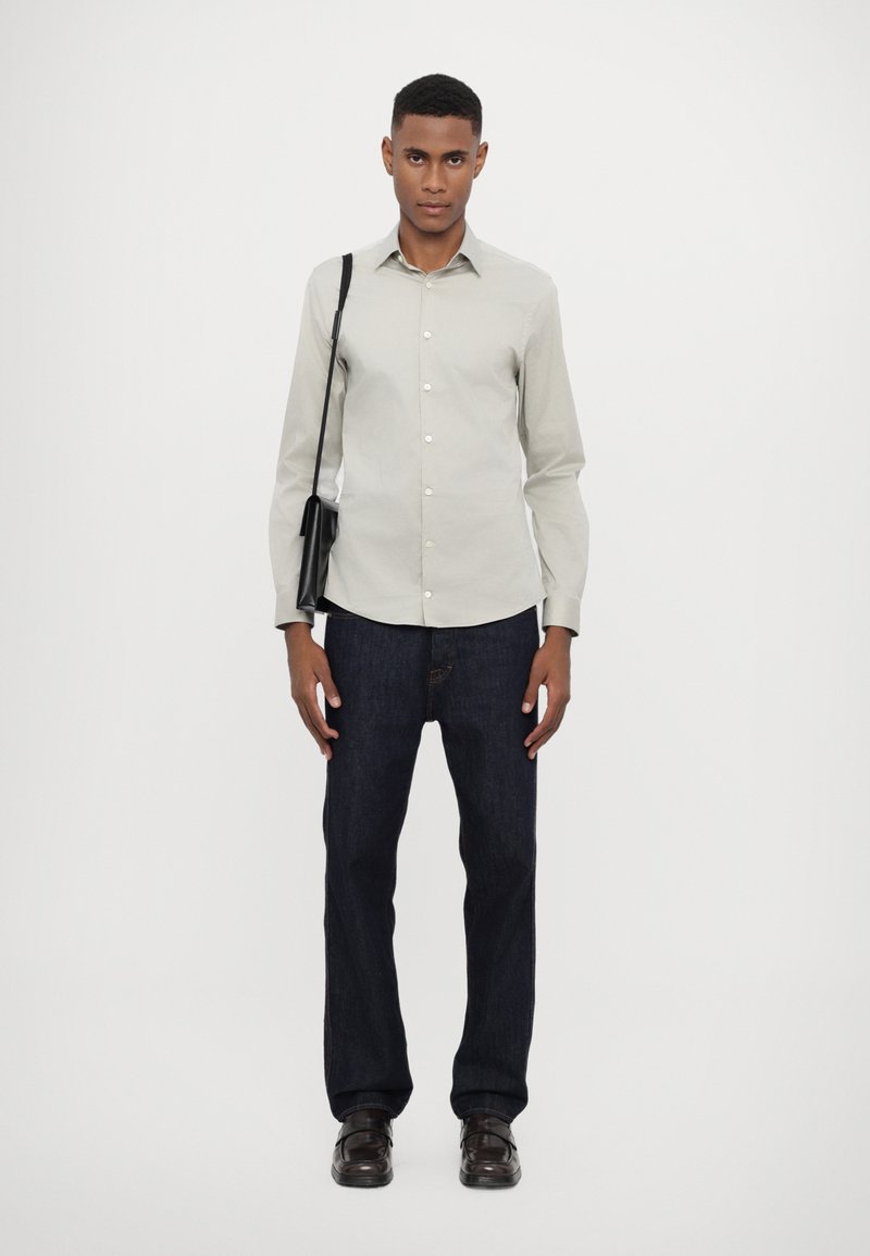 Chemise boutonnière gris clair, jean bleu foncé, sac à bandoulière noir et chaussures marron, se tenant contre un fond blanc.