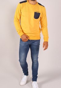 Maglione giallo a maniche lunghe con spalle testurizzate e taschino blu navy sul petto, abbinato a jeans blu attillati e sneakers bianche.