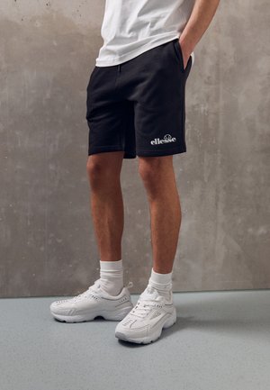 Man draagt zwarte ellesse short, witte sokken, witte sneakers en een wit shirt tegen een grijze, gestructureerde muur en vloer.