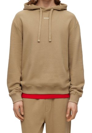 Beige-huppari, jossa nyörit ja "HUGO"-logo rinnassa, käytetty punaisen paidan päällä, yhdistetty beigen värisiin housuihin.