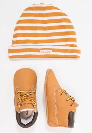 Stribet orange og hvid babyhue over et par brune babylædersko med gule og røde snørebånd, alt lagt fladt på en hvid baggrund.