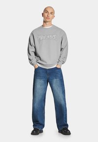 Szara bluza z białymi grafikami, noszona na białej koszuli. W połączeniu z jasnoniebieskimi luźnymi dżinsami i czarnymi butami, stojąca w neutralnej pozie.