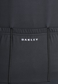 Musta urheilu paita, jossa on sileä pinta, ja jossa on heijastava "OAKLEY" -logo valkoisena sekä erottuvat ompeluyksityiskohdat.