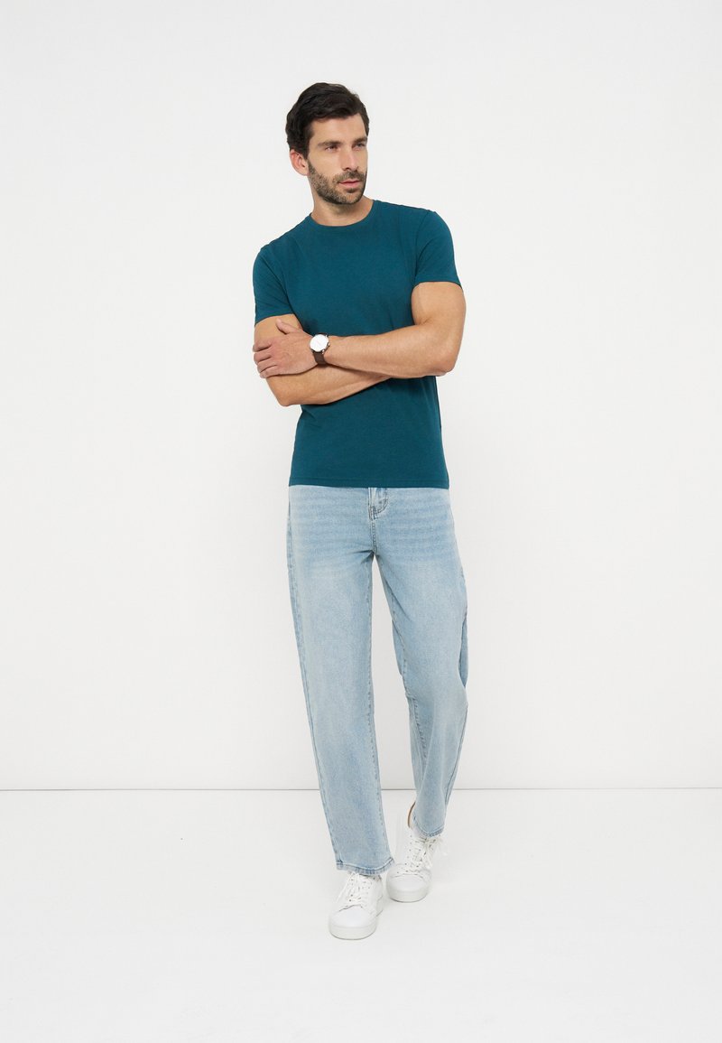 Mand med mørkt hår og skæg står med armene over kors iført en petroleumblå t-shirt, lyseblå jeans, hvide sneakers og et armbåndsur mod en hvid baggrund.