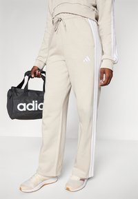 Bežās sporta biksēs ar baltām svītrām, elastīga josta, viegls audums un Adidas logotips. Melns sporta soma turēta vienā rokā.