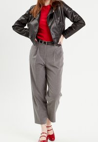 Sort lædermotorcykeljakke, rød skjorte, grå plisserede bukser og skinnende røde sko med ankelremme. Minimalistisk detaljering, glat finish.