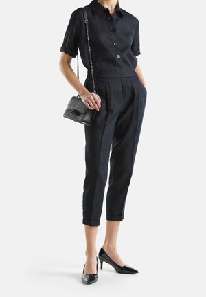 Frau trägt ein schwarzes, kurzärmeliges Hemd mit Knöpfen, zugeschnittene, verkürzte Hose, schwarze High Heels, eine Halskette als Choker und hält eine kleine schwarze Handtasche.