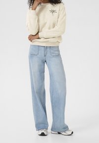 Sweatshirt crème avec broderie de palmier, associé à un jean large bleu clair. Mannequin portant des baskets blanches et noires.