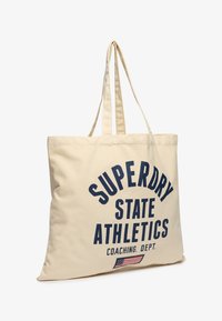 Beige canvas draagtas met lange hengsels, met grote blauwe tekst: "SUPERDRY STATE ATHLETICS COACHING DEPT." en een kleine Amerikaanse vlag graphic.
