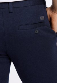 Mörkblå skräddarsydda byxor i texturerat tyg, med en bakficka med knappstängning och en liten märkesetikett.