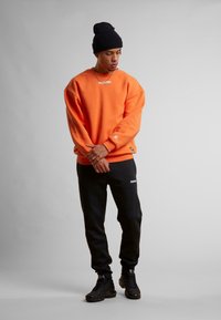 Oransje oversized hettegenser med et hvitt logo, kombinert med svarte joggebukser med et hvitt merkenavn, svarte joggesko og en svart lue.