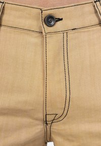Un pantalon en denim beige présente une fermeture à bouton à l'avant et des coutures et poches en contraste noir. Texture lisse, sans embellissements visibles.