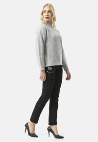 Vrouw met lang blond haar, gekleed in een grijze gebreide trui, zwarte slanke broek met ritsdetails en zwarte hoge hakken, kijkt naar links.