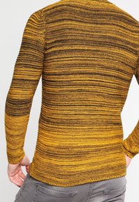 Svetr s dlouhým rukávem se vodorovnými pruhy v různých odstínech hořčicové a hnědé barvy, vyrobený z texturovaného pleteného materiálu.